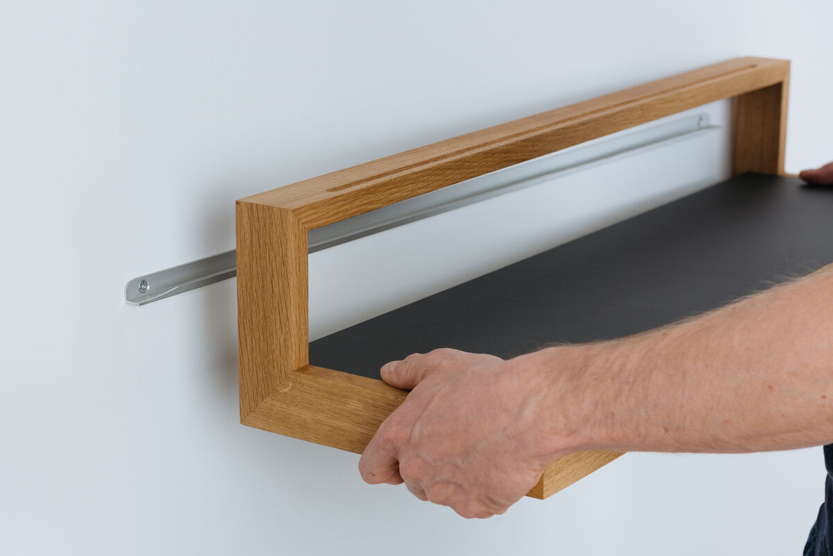 Ein anthrazitfarbenes Ablagebord mit einem hellbraunen Holzrahmen wird an einer Wand montiert. Zwei Hände sind dabei, es zu fixieren.