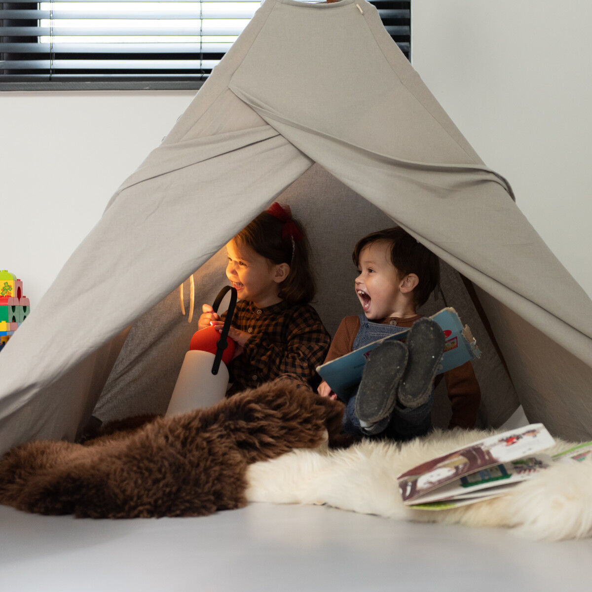 Zwei Kinder sitzen in einem beigen Zelt in einem gemütlichen Raum. Sie halten die »Guidelight« Lampe, die ein warmes Licht ausstrahlt, und lesen ein Buch zusammen. Die Umgebung ist kindgerecht dekoriert mit weichen Teppichen und Kissen.