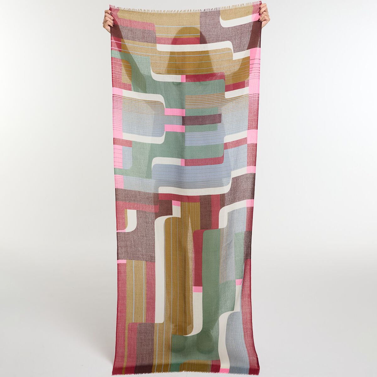 Ein langer, rechteckiger Wollschal im Farbton Khaki mit abstraktem, geometrischem Muster in Rosa, Hellblau, Beige, Braun und Weiß. Der Schal wird von einer Person mit ausgestreckten Armen vor neutralem Hintergrund präsentiert.