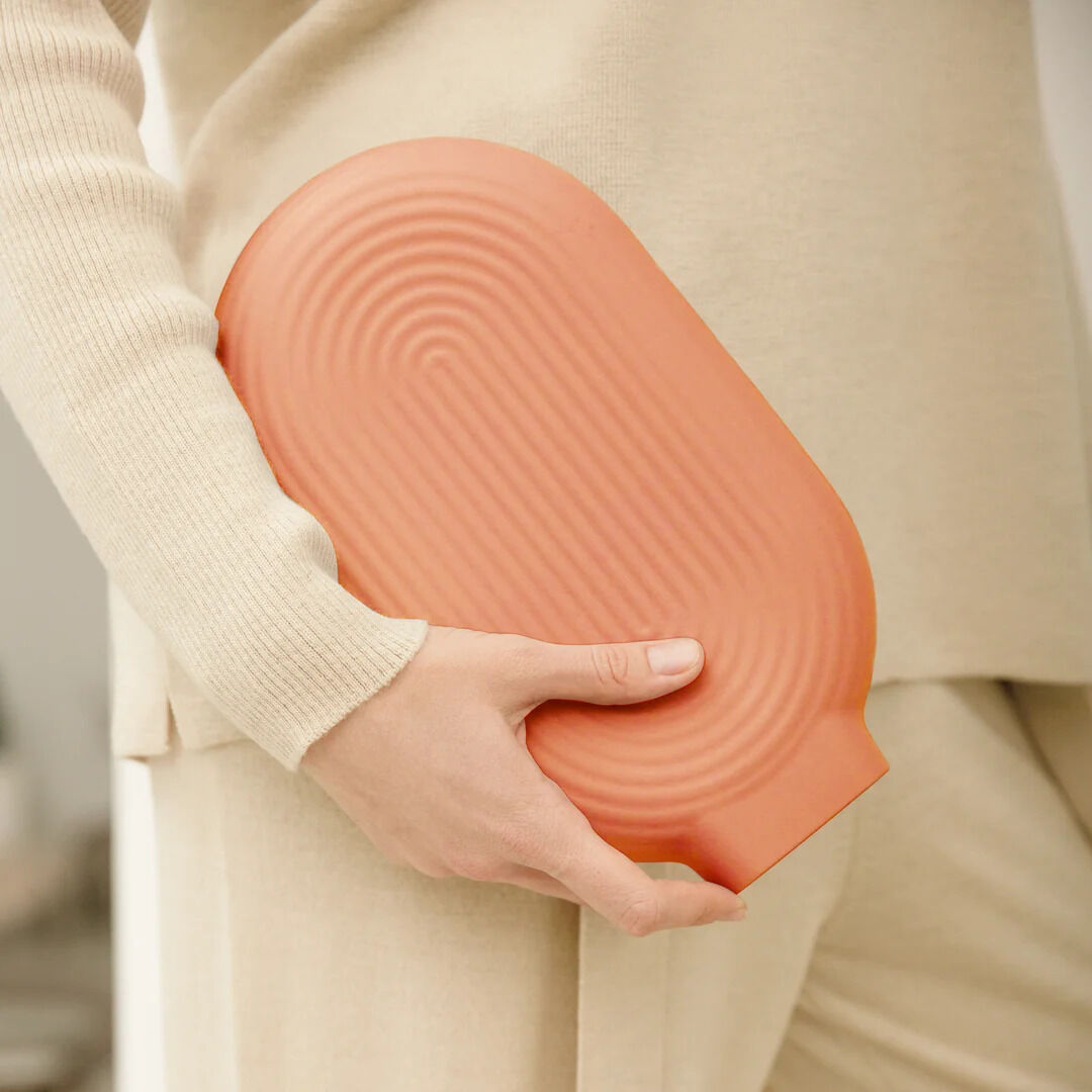 Nahaufnahme einer person in cremefarbener Kleidung, die eine korallenfarbene, wellenförmig strukturierte VAERM elektrische Wärmflasche Coralle aus Silikon im Soft-Touch-Material seitlich am Körper hält. Das Ambiente wirkt gemütlich, modern und einladend.