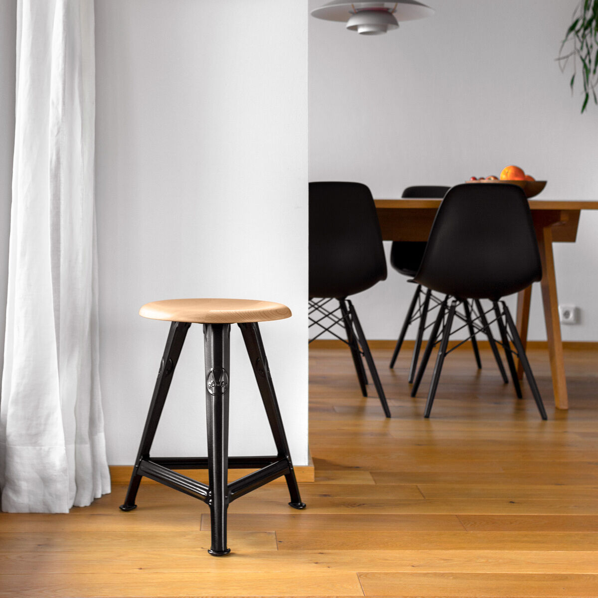 Der Rowac-Schemel »Eiche Mattschwarz« steht in einem minimalistisch eingerichteten Raum. Er hat eine Sitzfläche aus Holz und eine schwarz matten Metallbasis. Die Einrichtung umfasst einen Esstisch mit schwarzen Stühlen, einen hölzernen Boden und weiße Wände mit schlichter Dekoration.