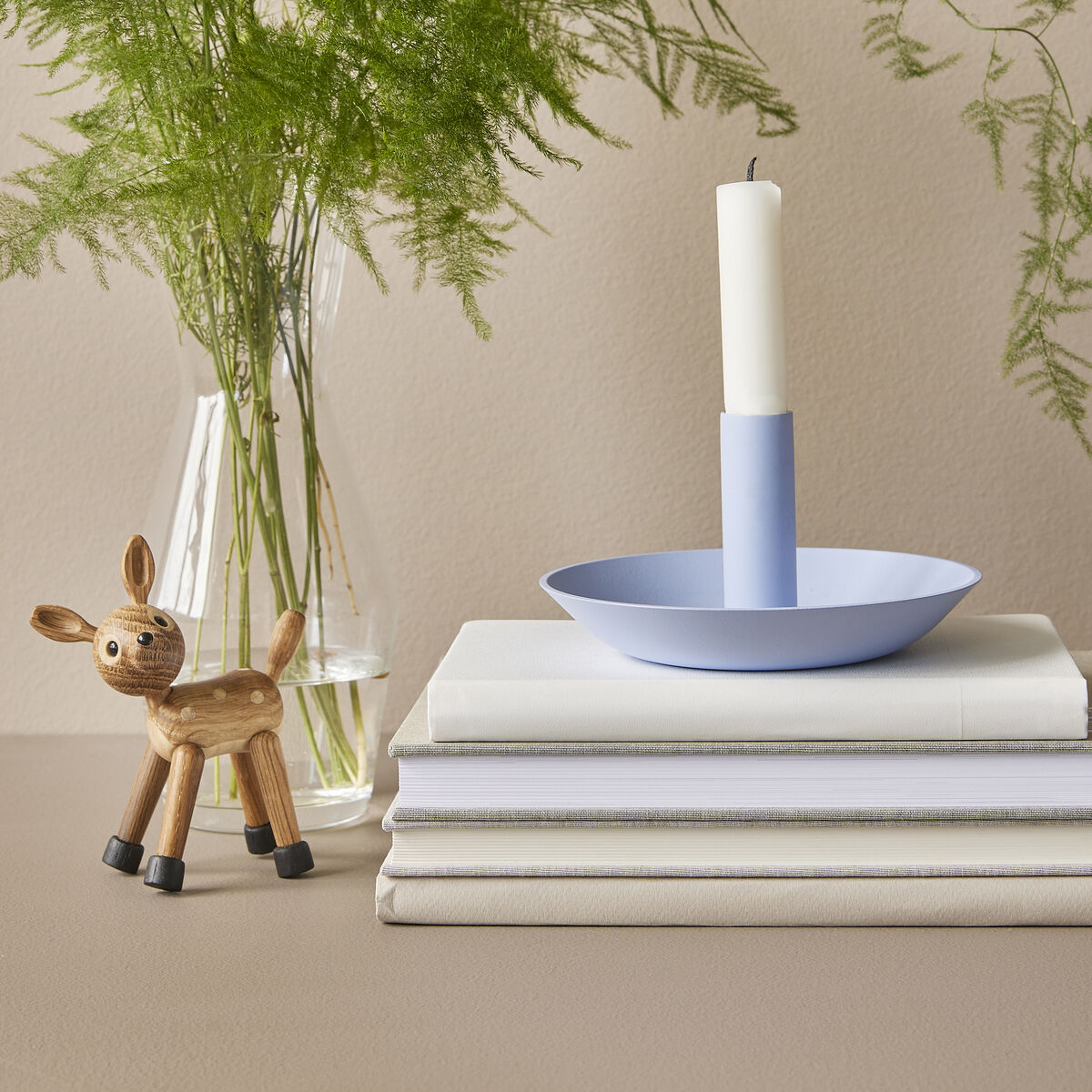 Holzfigur eines kleinen Tieres, arrangiert neben einer Glassvase mit grünem Farn, einem hellblauen Kerzenhalter mit weißer Kerze und drei aufeinander gestapelten Büchern auf einer beigen Oberfläche, alles im minimalistischen und hellen Einrichtungsstil fotografiert.