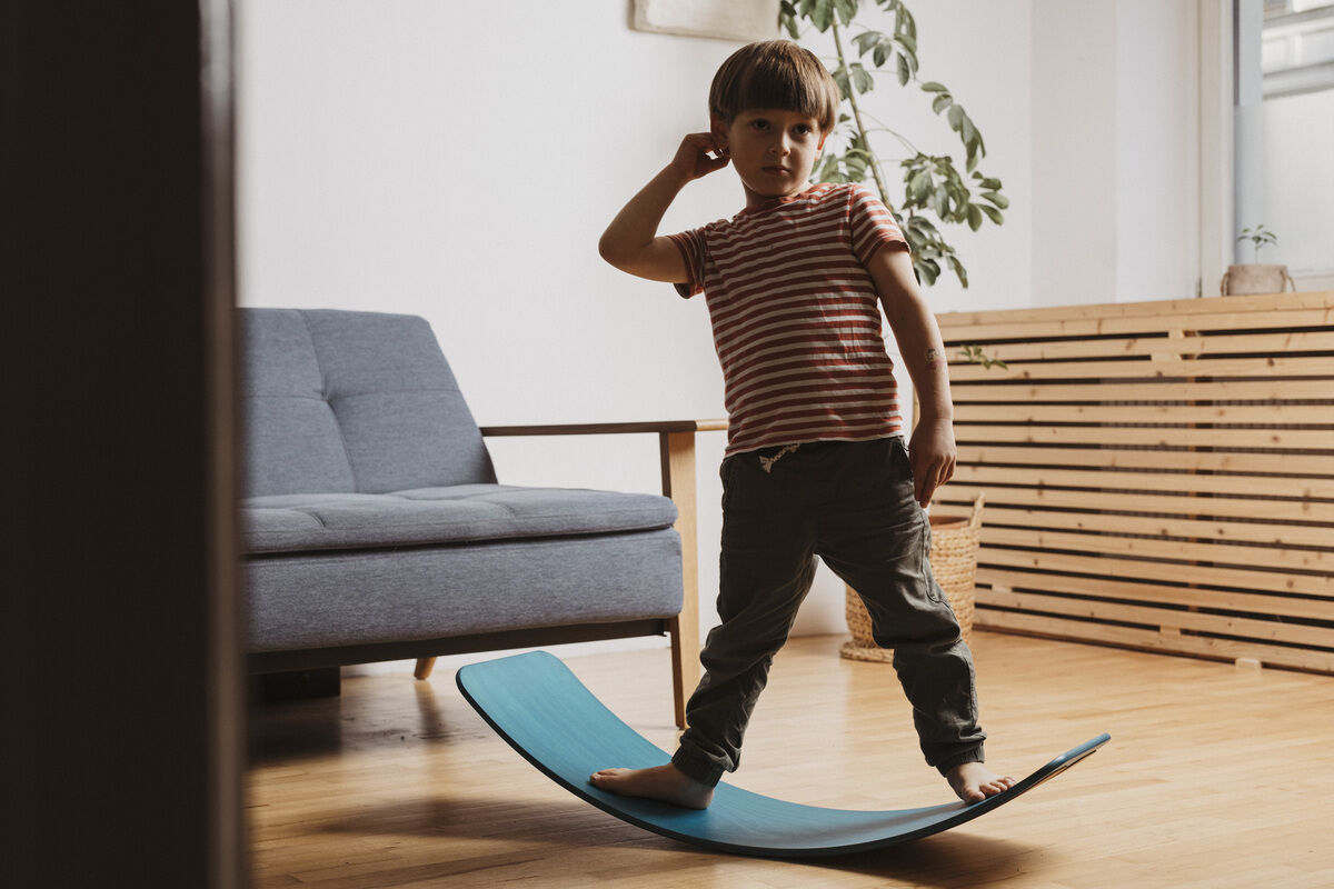 Ein Kind balanciert auf einem mintfarbenen Balance Board im Wohnzimmer. Im Hintergrund steht ein graues Sofa und eine Pflanze.