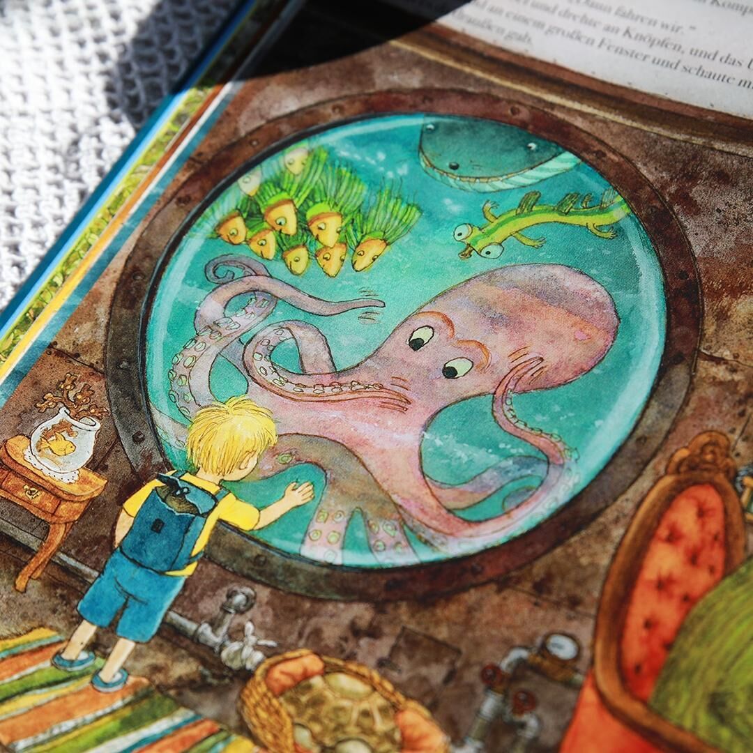Eine farbenfrohe Illustration aus einem Bilderbuch zeigt einen Jungen mit einem Rucksack, der durch ein großes Bullauge einen Oktopus und mehrere Fische beobachtet. Die Szene spielt in einem gemütlichen Raum mit Holzmöbeln und Teppichen. Der Oktopus und die Fische scheinen freundlich und neugierig.