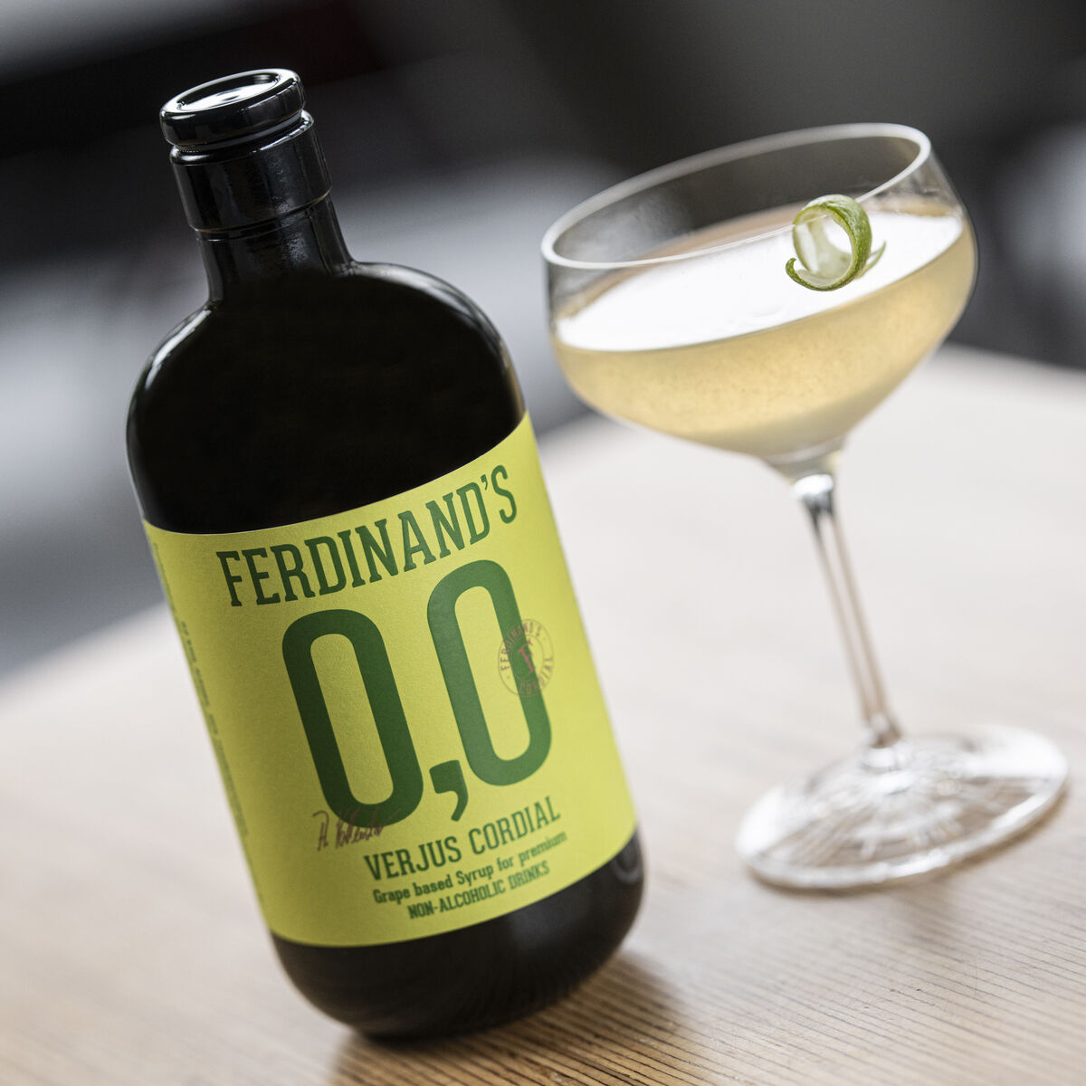 Eine grüne Flasche Ferdinands 0,0 Verjus Cordial steht auf einem Holztisch neben einem Cocktailglas mit einer hellem, leicht gelblichen Flüssigkeit, dekoriert mit einer dünnen Limettenspirale. Im Hintergrund sind unscharfe Möbel erkennbar.