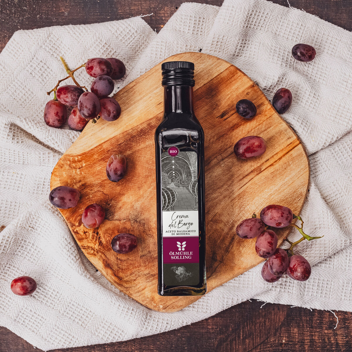 Eine Flasche dunkler Balsamico-Essig mit edlem Etikett steht auf einem runden Holzbrett, dekoriert mit frischen roten Weintrauben und einem weißen Stofftuch auf einem Holztisch.