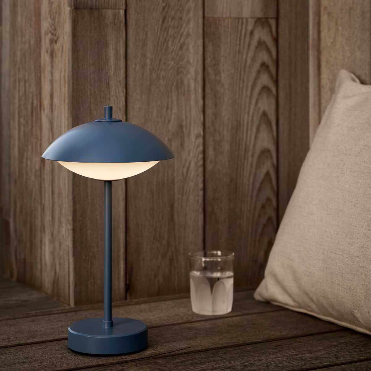 Tragbare Lampe »Clam™« von Fritz Hansen in mattem Blau auf hölzerner Sitzfläche vor einer rustikalen, vertikal getäfelten Holzvertäfelung. Daneben stehen ein Glas mit Wasser und ein helles Kissen, wodurch eine gemütliche, minimalistische Atmosphäre entsteht.