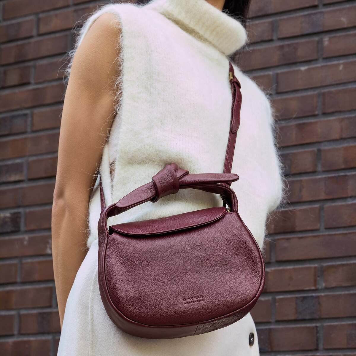 Eine Frau trägt eine weinrote Umhängetasche mit Knotendetail aus Lederoptik der Marke »Coco Croissant«. Sie trägt ein ärmelloses cremeweißes Outfit und steht vor einer braunen Backsteinwand. Die Tasche wirkt modern und elegant.