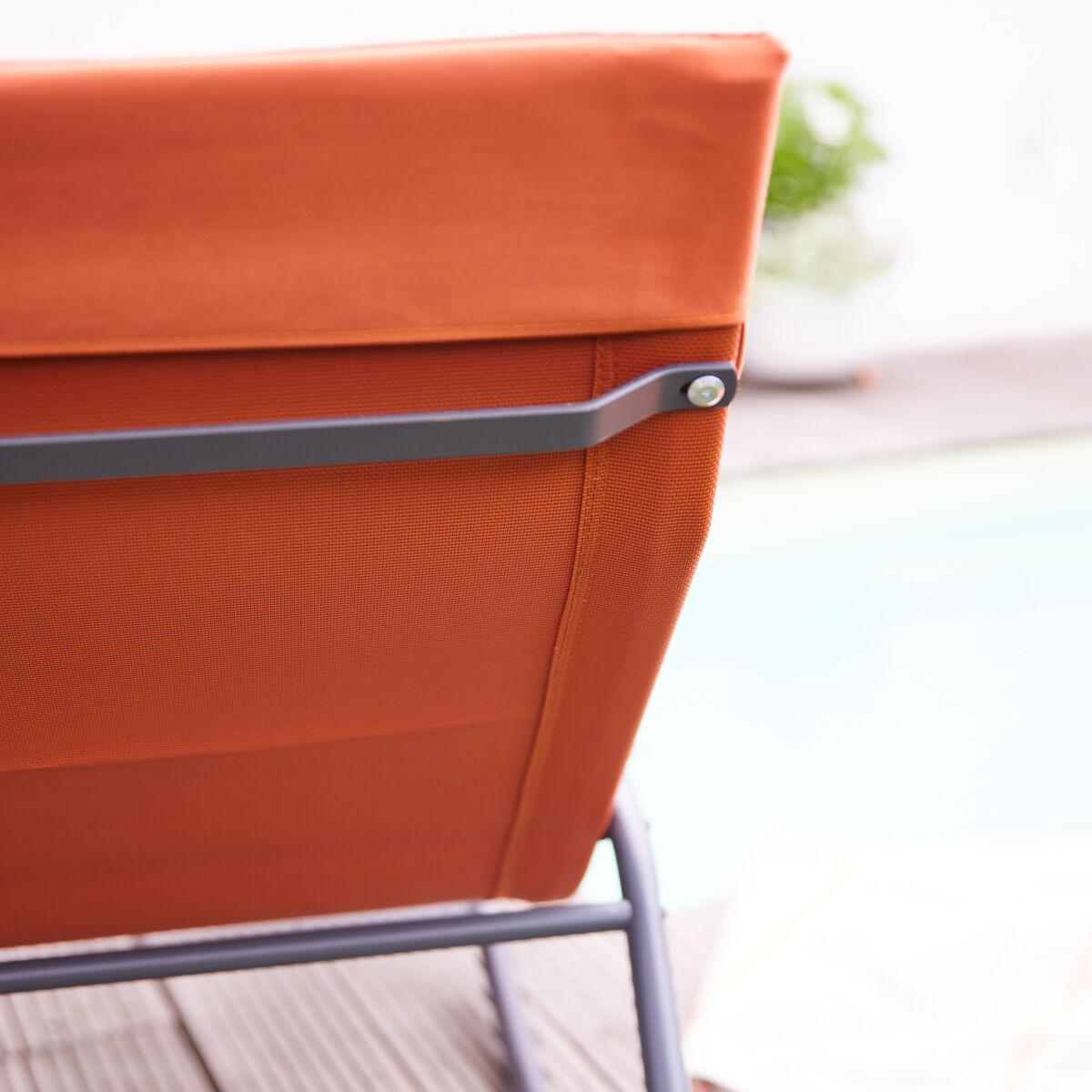 Nahaufnahme einer orangefarbenen Swingliege »Aliano« aus Metall und Stoff auf einer Holzterrasse. Im Hintergrund sind unscharf eine Poolkante und ein Handtuch zu erkennen. Die Szene vermittelt ein sommerliches, entspannendes Ambiente im Freien.