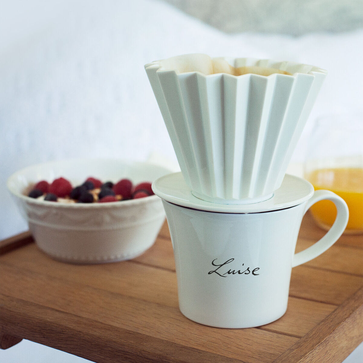 Eine Frühstückstasse »Berlin« von KPM mit der Schriftart Sententia. Die Tasse steht auf einem Holztisch neben einer Schale mit Obst und einem Glas Orangensaft. Oben auf der Tasse sitzt ein Filter.