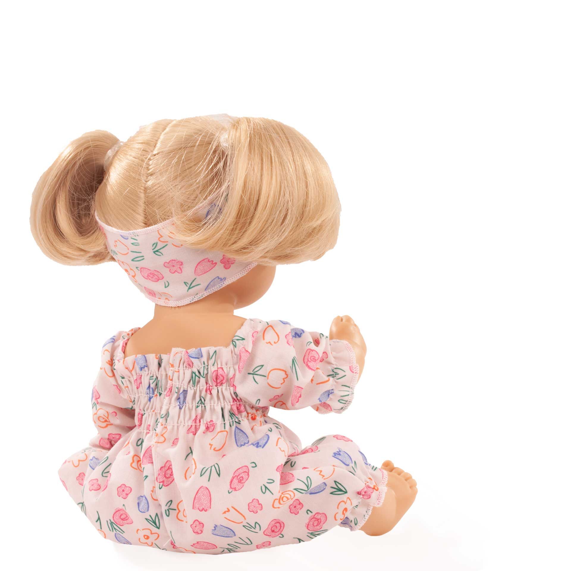 Eine Badepuppe aus der Serie Sleepy Aquini Girl Bliblablume sitzt aufrecht mit dem Rücken zum Betrachter. Sie trägt einen einteiligen Schlafanzug mit buntem Blumenmuster und ein passendes Stirnband. Die Puppe hat kurze, gebundene Haare und ist aus weichem Material gefertigt. Das Design ist kindgerecht und auf kleine Kinder zugeschnitten.