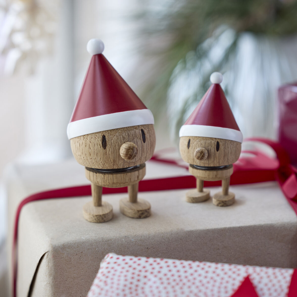 Zwei Hoptimist Elf Bumble Figuren aus Eiche mit roten Zipfelmützen stehen auf weihnachtlich verpackten Geschenken, verziert mit roten Bändern. Im Hintergrund ist unscharf Tannengrün zu sehen. Die Szene wirkt freundlich und festlich dekoriert.