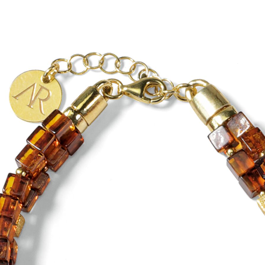 Das Bild zeigt ein elegantes Armband der Marke Ars mundi, Modell Annabele. Das Schmuckstück besteht aus kleinen kubischen Bernsteinperlen in warmem Braun, die in drei Reihen angeordnet sind. Die Verschlüsse und Kettchen sind vergoldetes 925er-Sterlingsilber, inklusive Anhänger mit dem eingravierten Logo 'AR'.