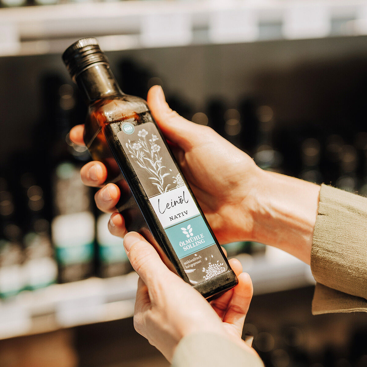 Detailaufnahme einer Hand, die eine Flasche Ölmühle Solling Leinöl NATIV hält. Das Etikett zeigt botanische Zeichnungen und Text. Im Hintergrund stehen weitere Flaschen unscharf im Regal. Die Szene ist freundlich beleuchtet und vermittelt einen natürlichen, hochwertigen Eindruck.