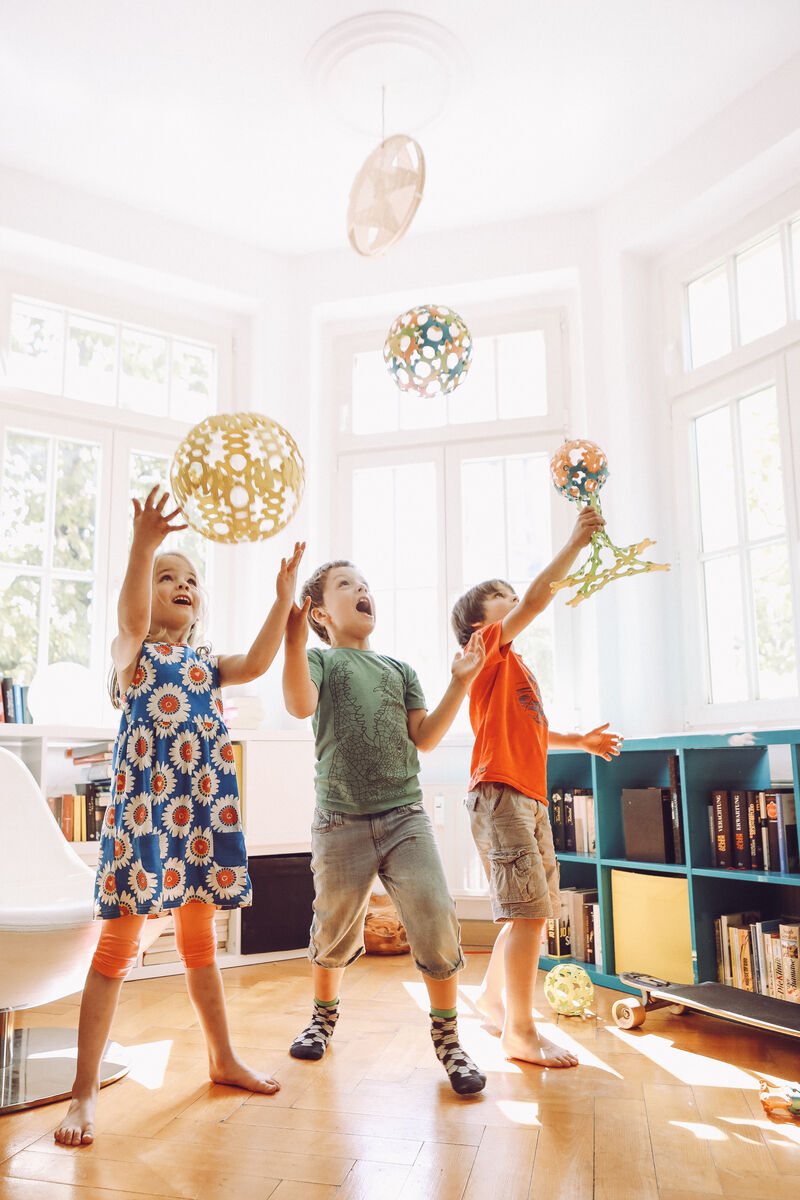 Drei Kinder spielen drinnen mit bunten, kugelförmigen Bauten aus dem Spiel »Binabo« 240 Teile. Sie lachen und heben die bunten Gebilde in die Luft. Im Hintergrund sind große Fenster und Regale mit Büchern zu sehen. Das Zimmer ist hell und freundlich gestaltet.