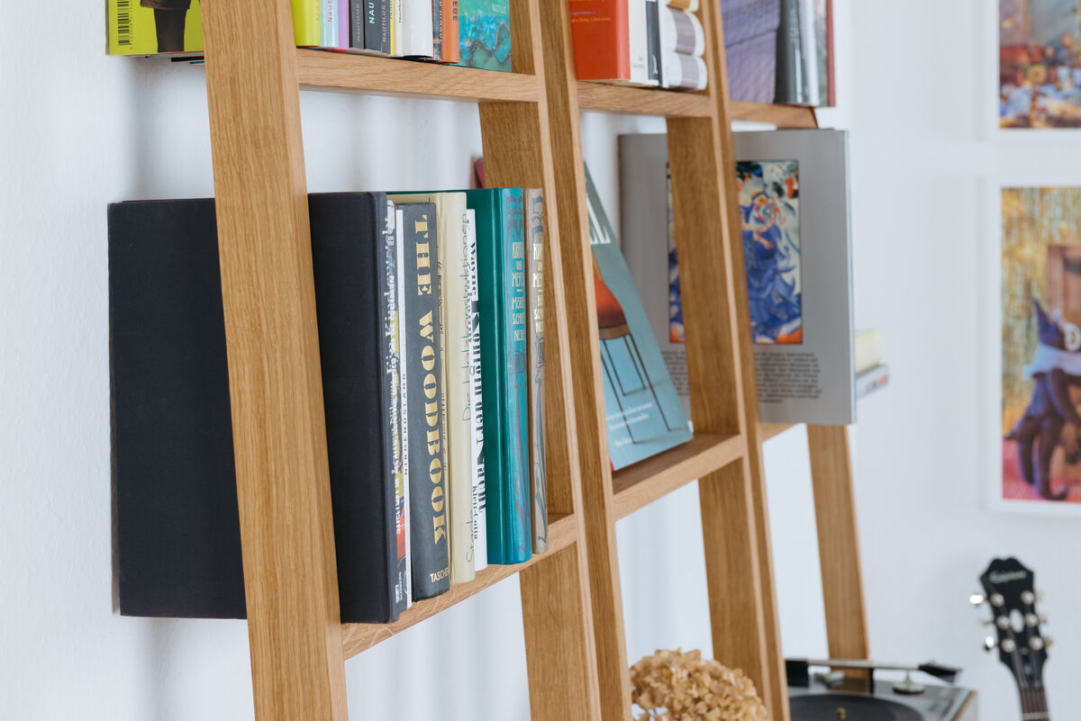 Ein Leiterregal namens »brecht« aus Holz, das an einer Wand aufgestellt ist. Das Regal ist mit mehreren Büchern und Dekorationselementen bestückt. Das Bild zeigt einen Ausschnitt des Regals im Kontext eines modern eingerichteten Raumes mit weiß gestrichener Wand.