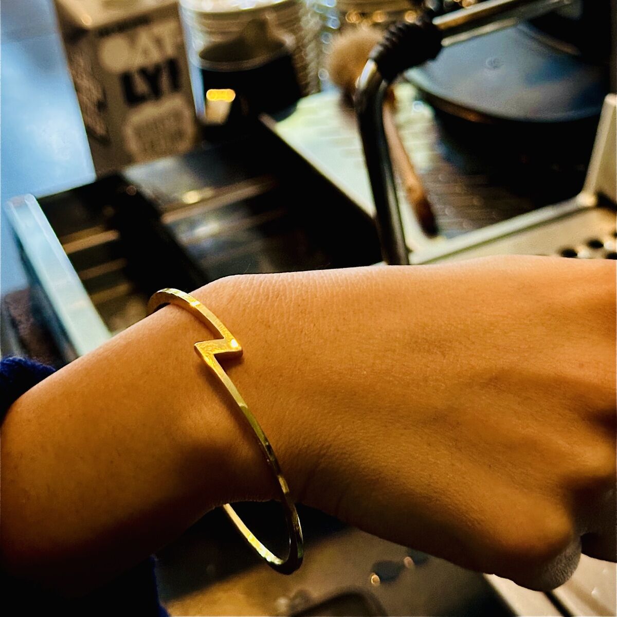 Ein goldplattierter Armreif der ZEIT Sonderedition von HML Berlin im Zickzack-Design schmückt ein Handgelenk. Im Hintergrund befinden sich eine Kaffeemaschine, Kaffeetassen und ein Tetrapak Oatly Barista Edition.
