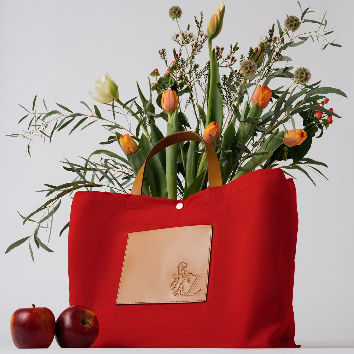 Eine rote Jubiläums-Markttasche mit Ledergriffen, gefüllt mit frischen Blumen, darunter Tulpen und Gräser. Vor der Tasche liegen zwei rote Äpfel.