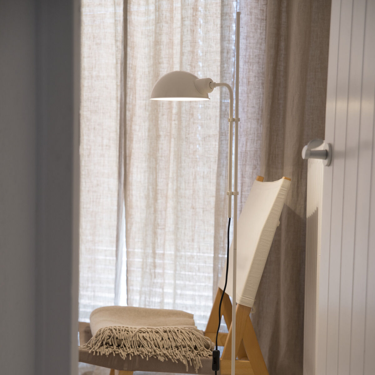 Eine moderne Stehleuchte »Funiculi« in einem minimalistischen Wohnzimmer. Die Lampe ist weiß und steht neben einem Stuhl aus Holz mit einem beigen Sitzkissen. Helle Vorhänge und ein Holzfußboden sind ebenfalls zu sehen.