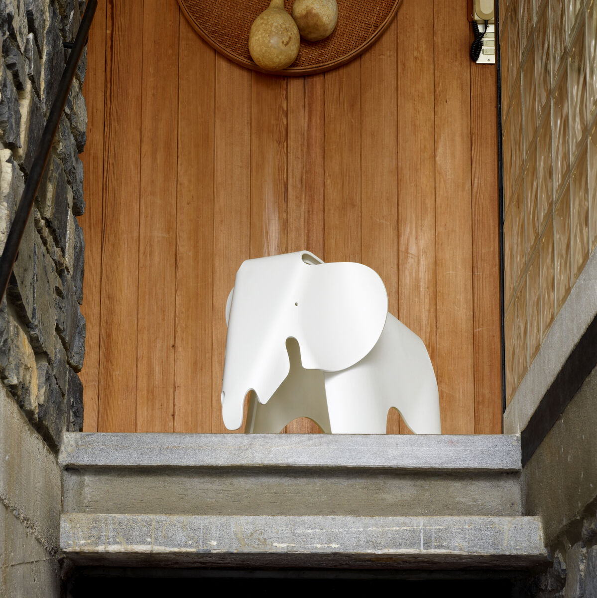Ein Kinderhocker in Form eines weißen Elefanten, bekannt als Eames Elephant, steht auf einer Steintreppe. Dahinter ist eine Holztür und eine Wanddekoration sichtbar. Die Umgebung besteht aus Steinmauern und modernen Designelementen.