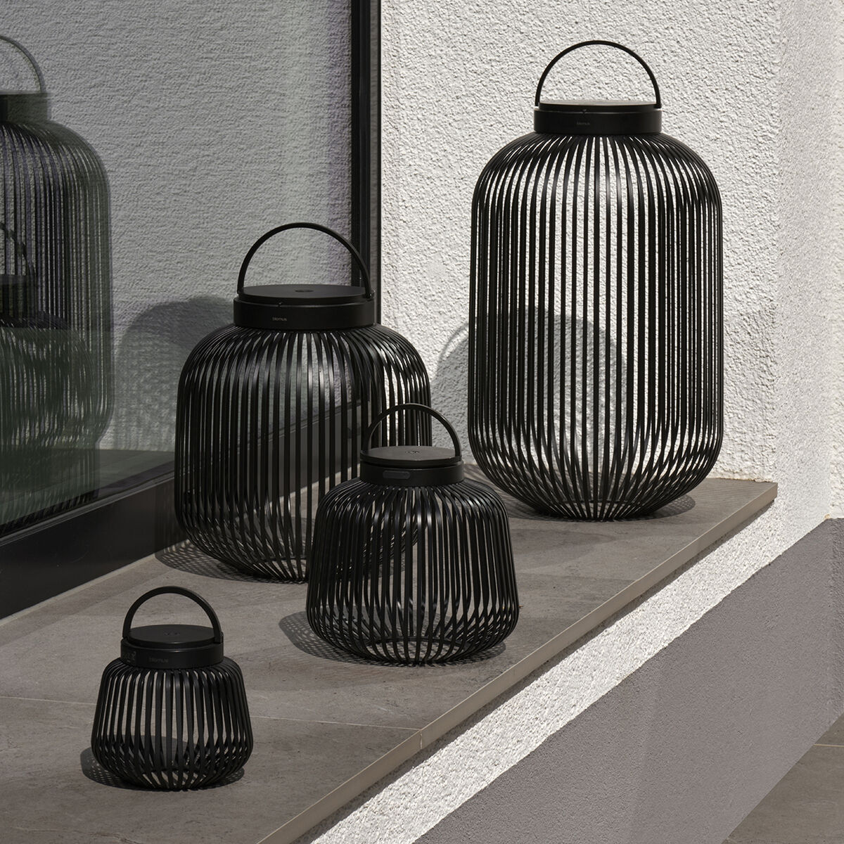 Vier LED-Leuchten der Marke blomus in Granite Grey stehen auf einem Sims vor einer Fensterfront. Die modernen, runden Leuchten mit engen, vertikal verlaufenden Streben aus Metall variieren in der Größe und wirken skulptural sowie dekorativ für drinnen und draußen.