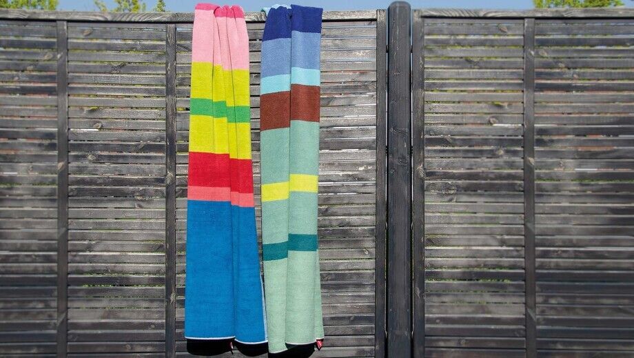 Zwei bunte Badetücher hängen an einem dunklen Holzzaun. Die Handtücher haben verschiedene Streifenmuster in den Farben Blau, Grün, Rot, Gelb, Rosa und Braun.