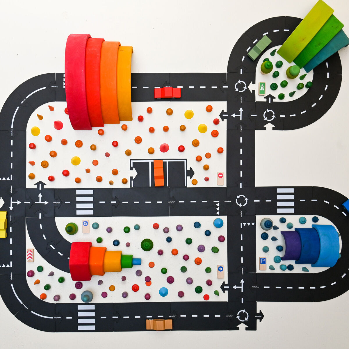 Obenansicht eines ausgebreiteten Straßensets mit aufgeklebten, schwarzen Straßenlinien, bunten Holzelementen, kleinen farbigen Autos und verschiedenen Bausteinen, arrangiert als Stadtlandschaft auf einem hellen Untergrund. Das Spielzeug eignet sich besonders für Kinder.
