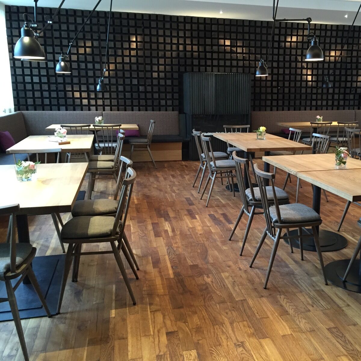 Ein modernes Restaurant mit Holzboden und Holztischen, die von Ercol-Stühlen in grau gebeizt umgeben sind. Die Stühle sind schlicht gestaltet und passen gut zum skandinavischen Design des Raumes. Mehrere Blumenarrangements stehen auf den Tischen als Dekoration.