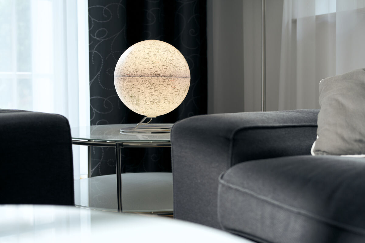 Ein Leuchtglobus von National Geographic mit dem Thema Mond, platziert auf einem Tisch in einem modernen Wohnzimmer. Der Globus leuchtet angenehm und erzeugt eine stimmungsvolle Beleuchtung. Die Umgebung ist in Schwarz- und Grautönen gehalten.
