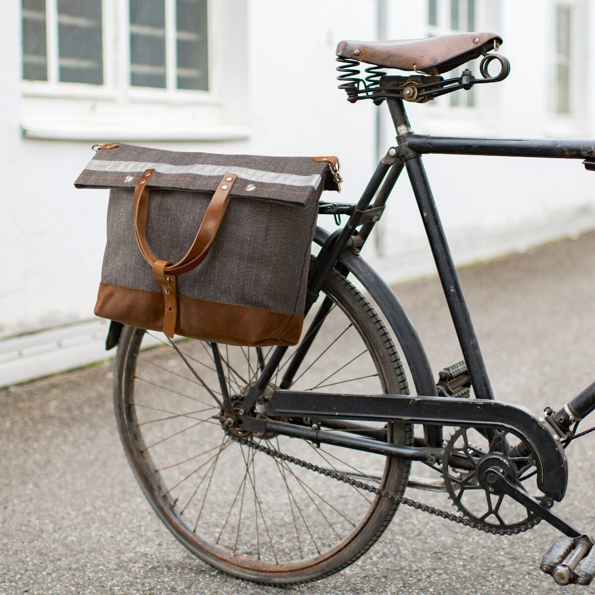 Fahrradtasche aus braunem und grauem Stoff, angebracht an einem schwarzen Fahrrad mit braunem Ledersattel, geparkt auf einem asphaltierten Gehweg vor einem weißen Gebäude.