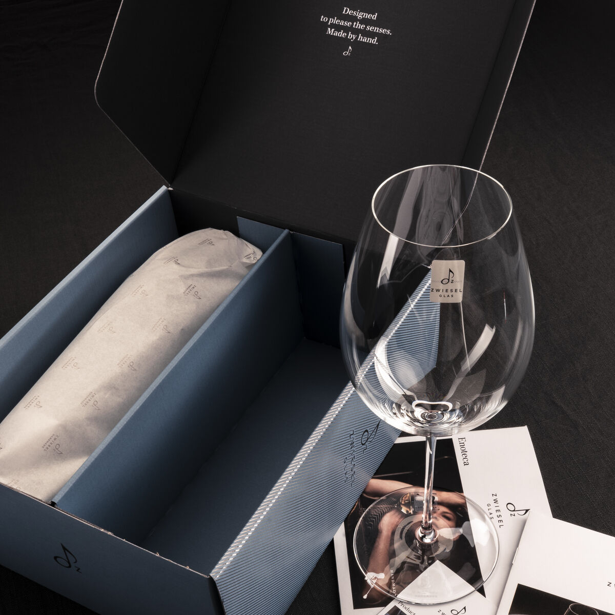 Ein Rotwein 2er-Set »Enoteca« bestehend aus einem eleganten Weinglas und einer stilvollen Geschenkverpackung. Das Set liegt auf einem dunkelgrauen Hintergrund mit einigen Prospekten daneben.