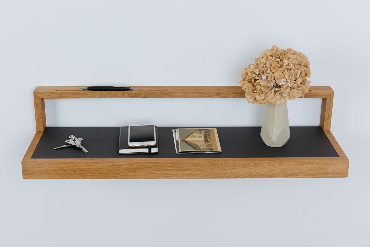 Ablagebord »buck« in Anthrazit mit einer Oberfläche aus Holz. Auf dem Regal befinden sich ein Schlüsselbund, ein Notizbuch, ein Buch, ein dekorativer Papierfächer und eine Vase mit getrockneten Blumen. Das Regal ist an einer weißen Wand befestigt.
