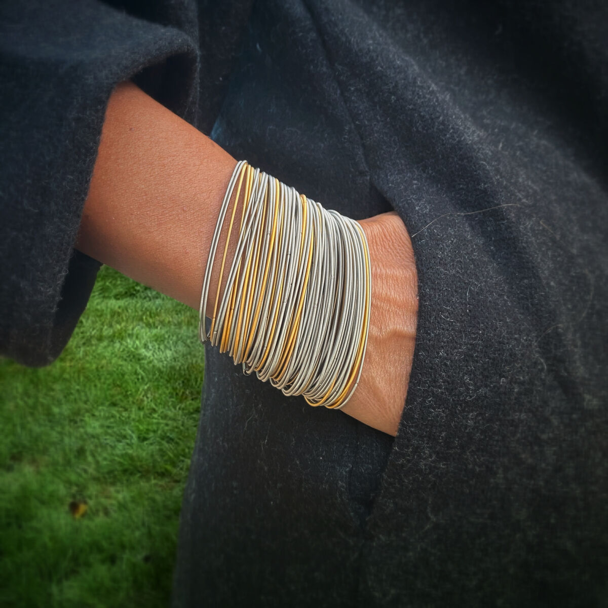 Ein modisches Armband aus einer Vielzahl von dünnen Drahtsträngen in Schwarz, Grau, Gold und Silber, getragen an einem Handgelenk in einer dunklen Jackentasche.