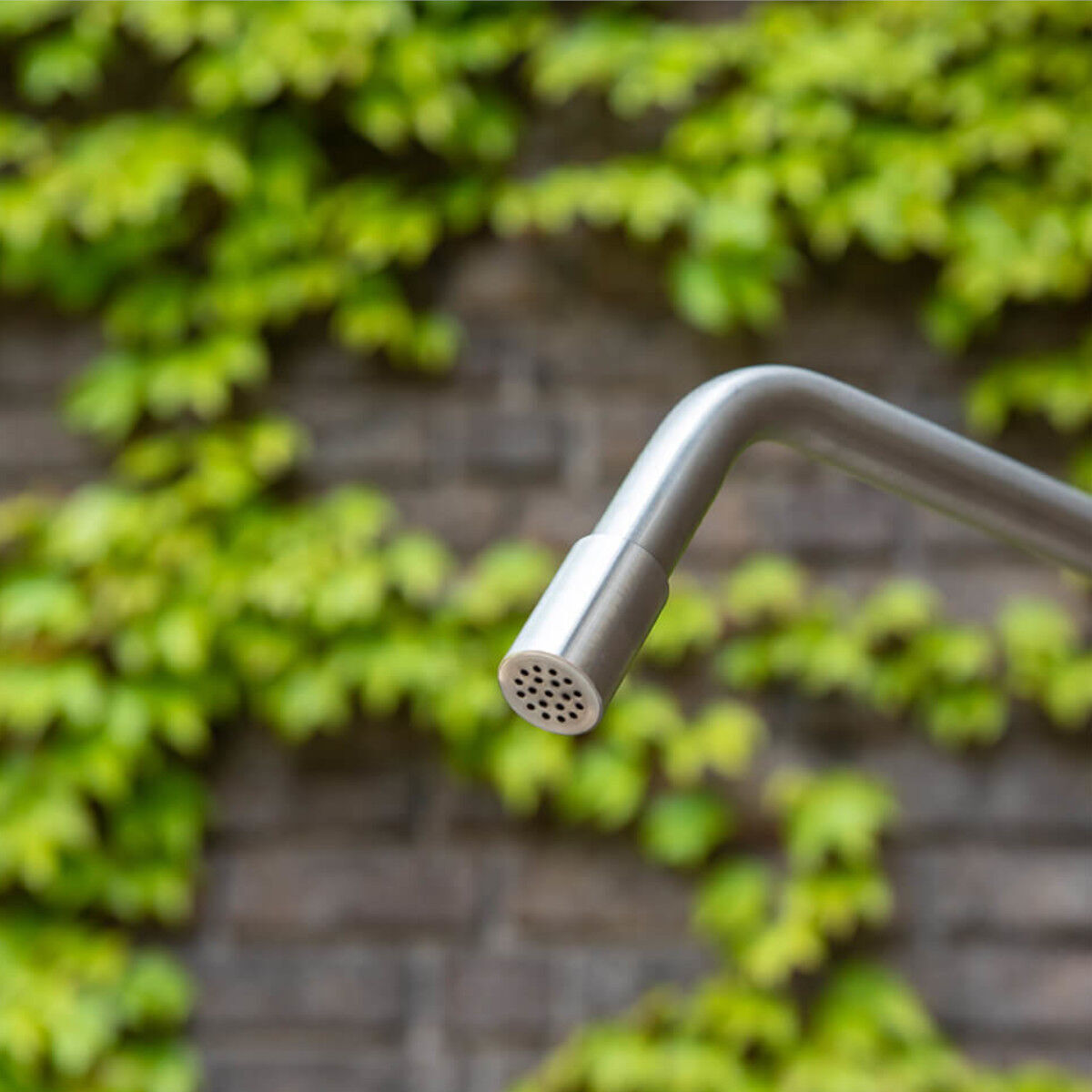 Nahaufnahme einer Gartendusche »Serpentine« aus Edelstahl vor einem Hintergrund von belaubten Pflanzen und einer Backsteinmauer.