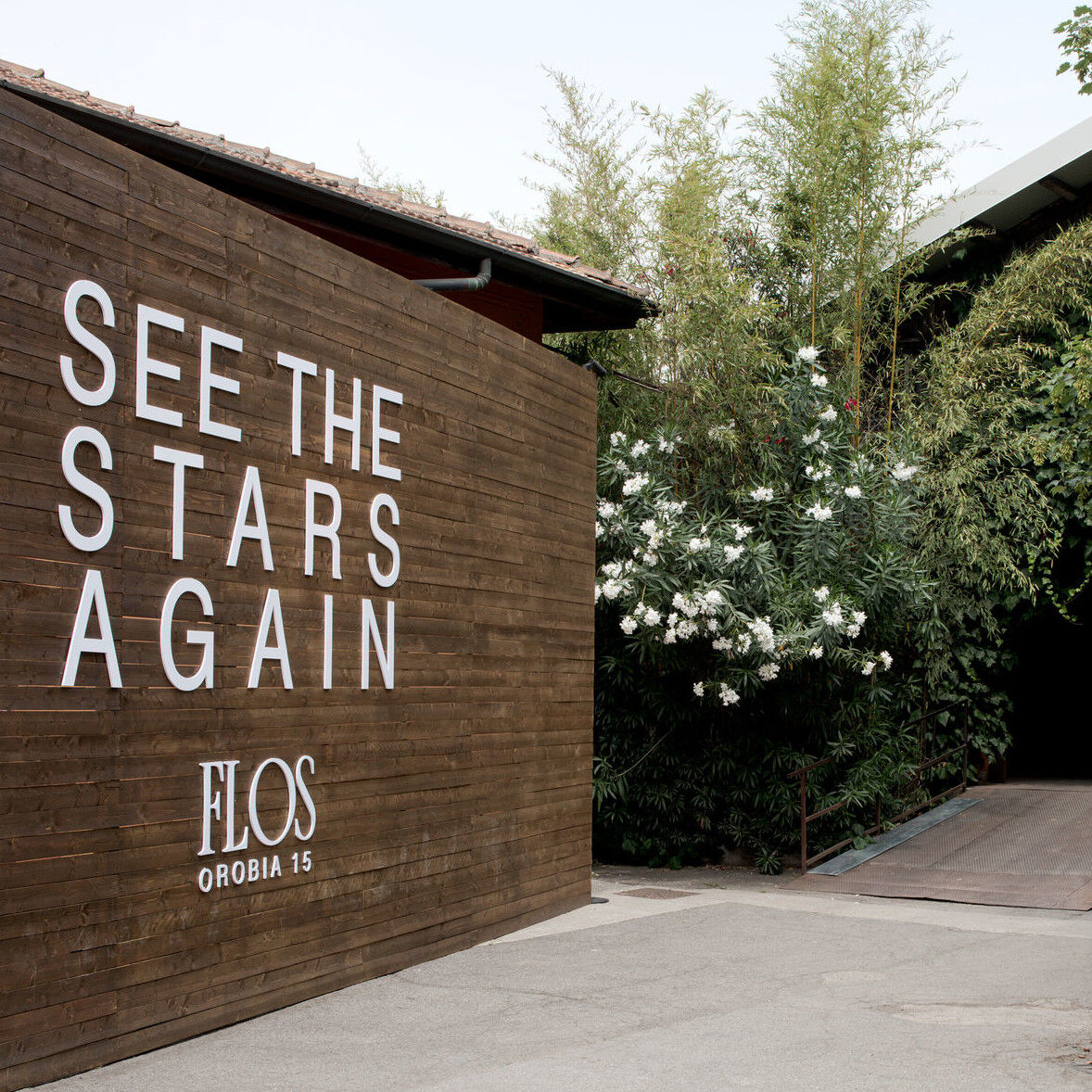 Holzwand mit weißer Aufschrift 'SEE THE STARS AGAIN' und 'FLOS OROBIA 15' neben grünen Büschen und Bäumen
