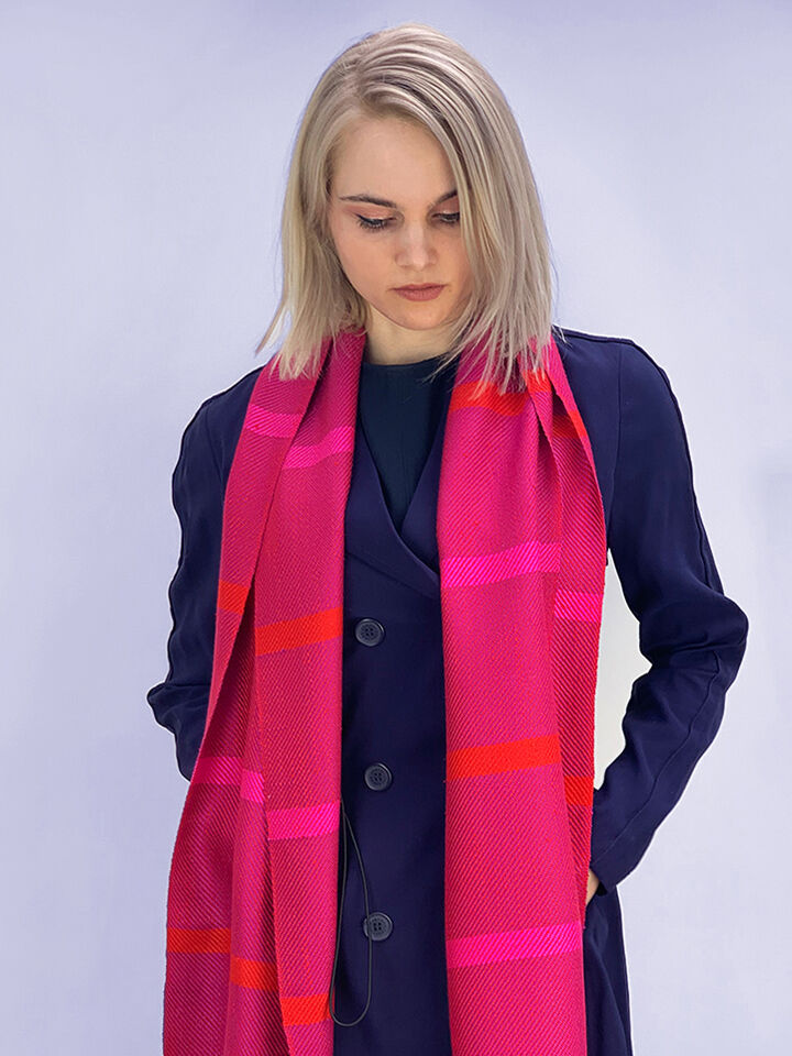 Eine Frau trägt einen langen gewebten Seidenschal in Fuchsia und Rot über einem dunkelblauen Mantel. Sie schaut nach unten und ihre Hände sind in den Taschen ihres Mantels vergraben. Der Hintergrund ist neutral und hell.