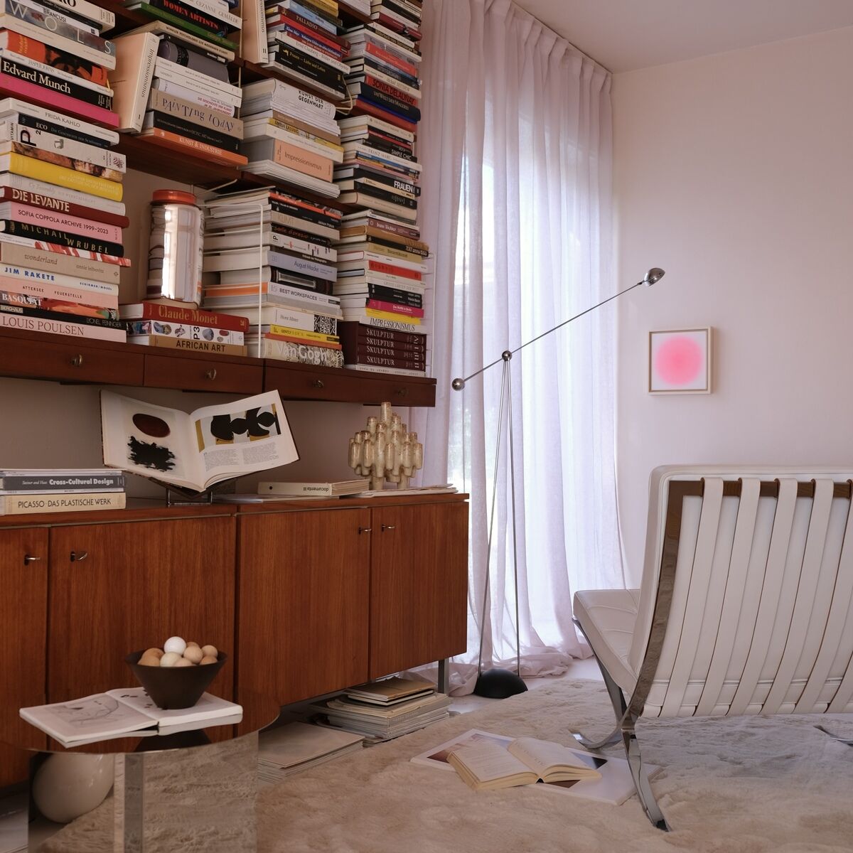 Ein modern eingerichtetes Wohnzimmer mit bodentiefen weißen Vorhängen, einem großen Holzsideboard und einem Bücherregal voller bunter Bücher. Im Vordergrund eine weiße Designer-Lounge-Stuhl und ein kleiner Tisch mit Dekoration. An der Wand hängt das quadratische Kunstwerk ZEIT-Sonderedition Sorbet »Pink«, das durch seine pastellrosa Farbgebung auffällt.