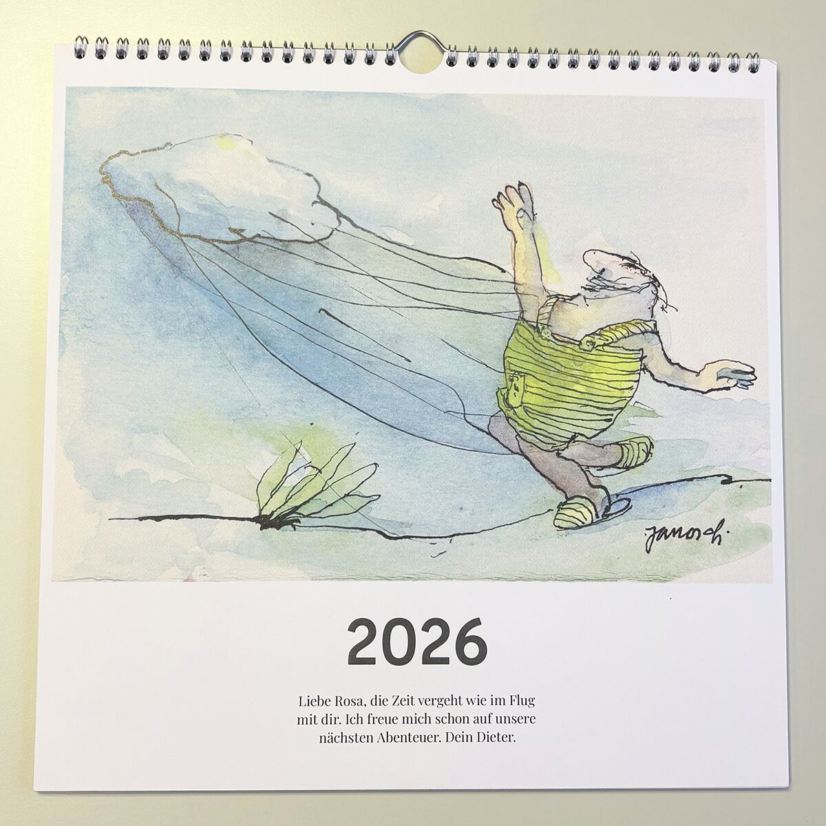Der Wondrak-Kalender 2026 zeigt ein humorvolles, gezeichnetes Kalenderblatt mit einer Cartoon-Figur in grüner Kleidung und einem bräunlichen Hut. Die Illustration ist in Aquarell- und Skizzenstil gehalten, darunter steht der Jahreszahl 2026 sowie ein kurzer, persönlicher Spruch. Oberhalb befindet sich eine silberne Spiralbindung. Das Design wirkt freundlich und künstlerisch. Ideal als Wandkalender für Zuhause oder das Büro.