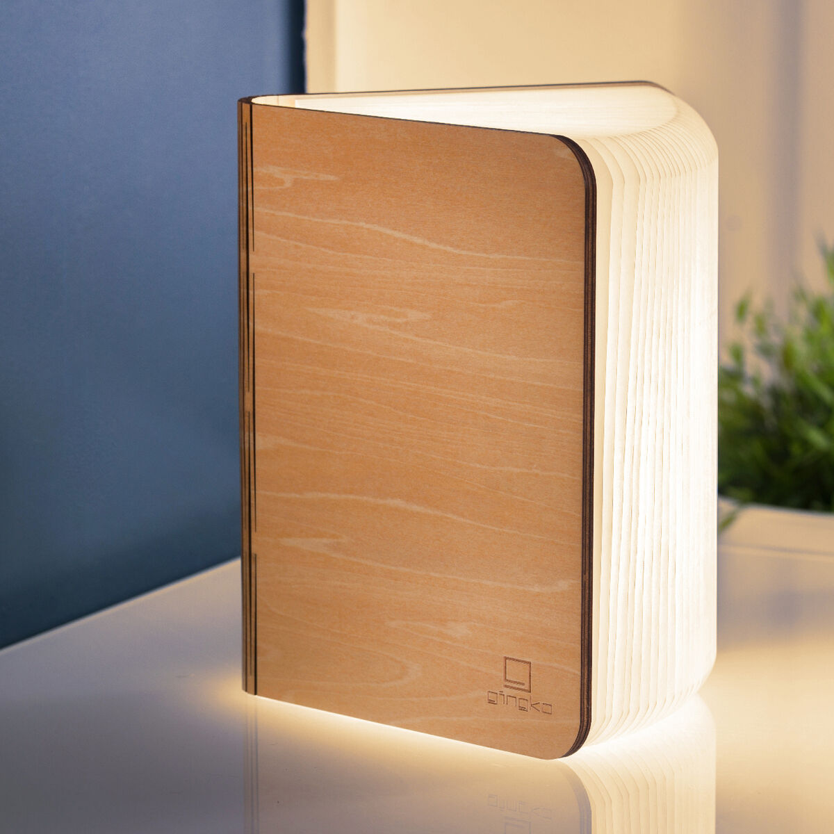 Ein Smart-Booklight in Ahorn-Holzoptik steht auf einem weißen Tisch, umgeben von Schreibutensilien und Pflanzen, und strahlt warmes Licht aus.
