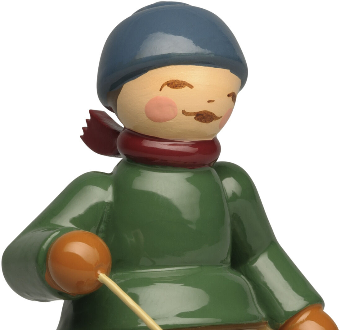 Detailreiche Holzfigur »Winterkinder - Junge auf Schlitten« von Wendt & Kühn. Ein Junge mit grüner Jacke, blauer Mütze und rotem Schal sitzt auf einem hellen Holzschlitten, hält zwei Stöcke und trägt schwarze Stiefel. Die Figur wirkt handbemalt und fröhlich.