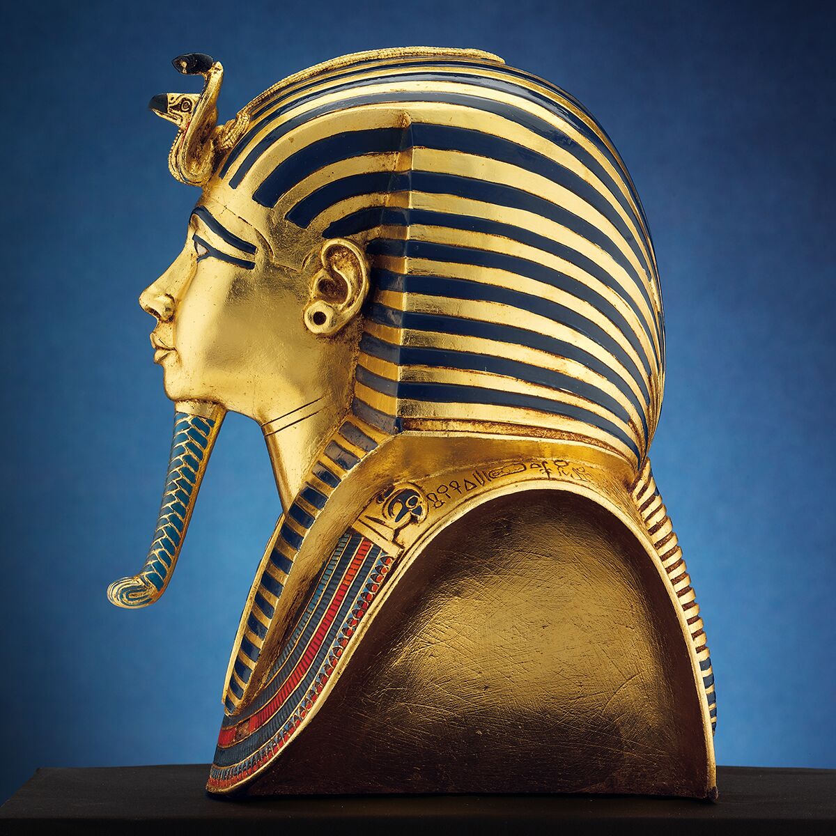 Seitliche Ansicht einer detailreichen Büste der berühmten Goldmaske des Tutanchamun. Das Kunstwerk besteht aus poliertem goldfarbenem Kunstguss mit blauen und roten Akzenten und ist auf einem schwarzen Sockel montiert. Hintergrund blau leuchtend, Exponat wirkt edel und wertvoll.