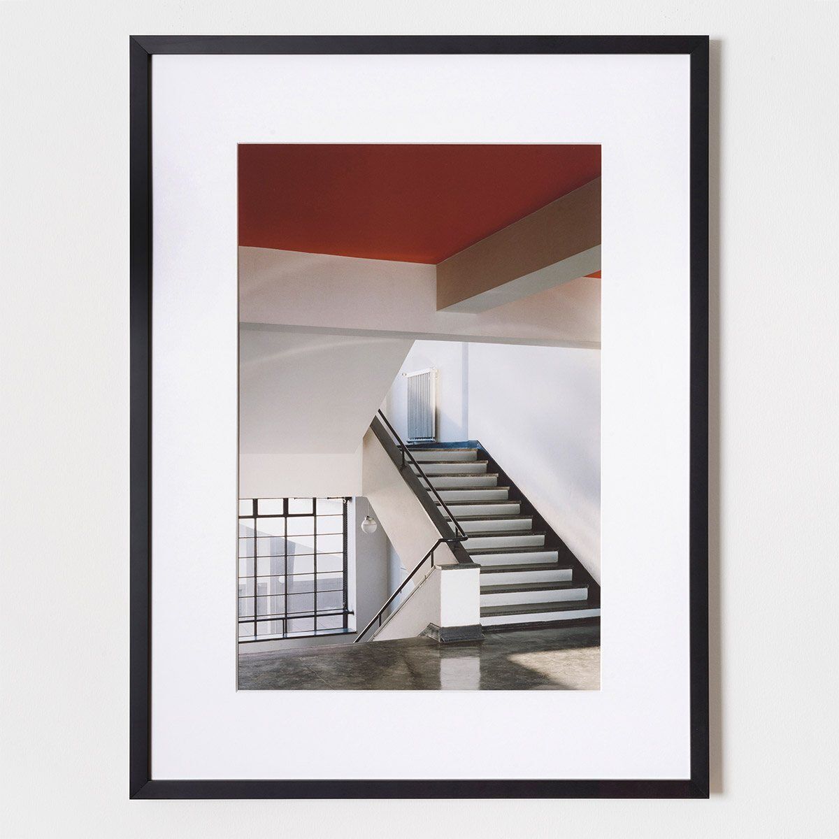 Fotografie von Klaus Kinold zeigt das Haupttreppenhaus im Bauhaus Dessau, entworfen von Walter Gropius. Die Aufnahme von 1992 betont die modernen geometrischen Linien und die Kontraste zwischen den Farben Weiß, Rot und Schwarz. Das Bild ist in einem schwarzen Rahmen gefasst.