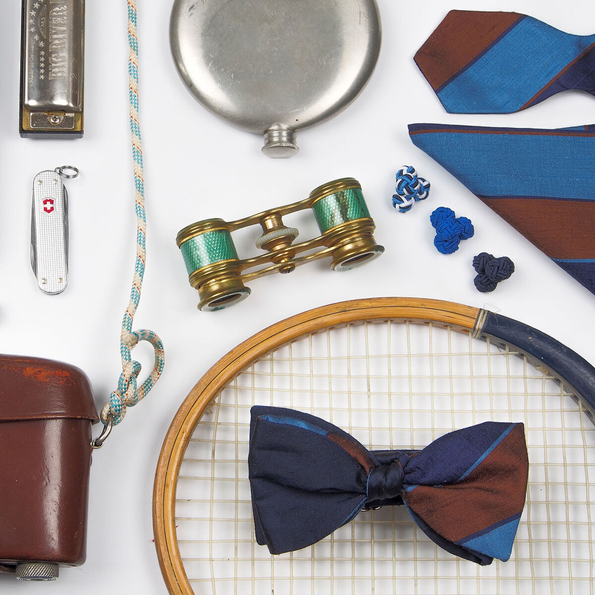 Das Accessoire-Set #007 Goldenstripe's Edition beinhaltet eine stilvolle Kombination von Accessoires wie eine blaue und braune Krawatte, eine dazu passende Fliege, retro Manschettenknöpfe, ein Schweizer Taschenmesser, ein Fernglas, eine silberne Flasche, ein Seil und ein Vintage-Tennisracket, alle auf einer weißen Oberfläche arrangiert.
