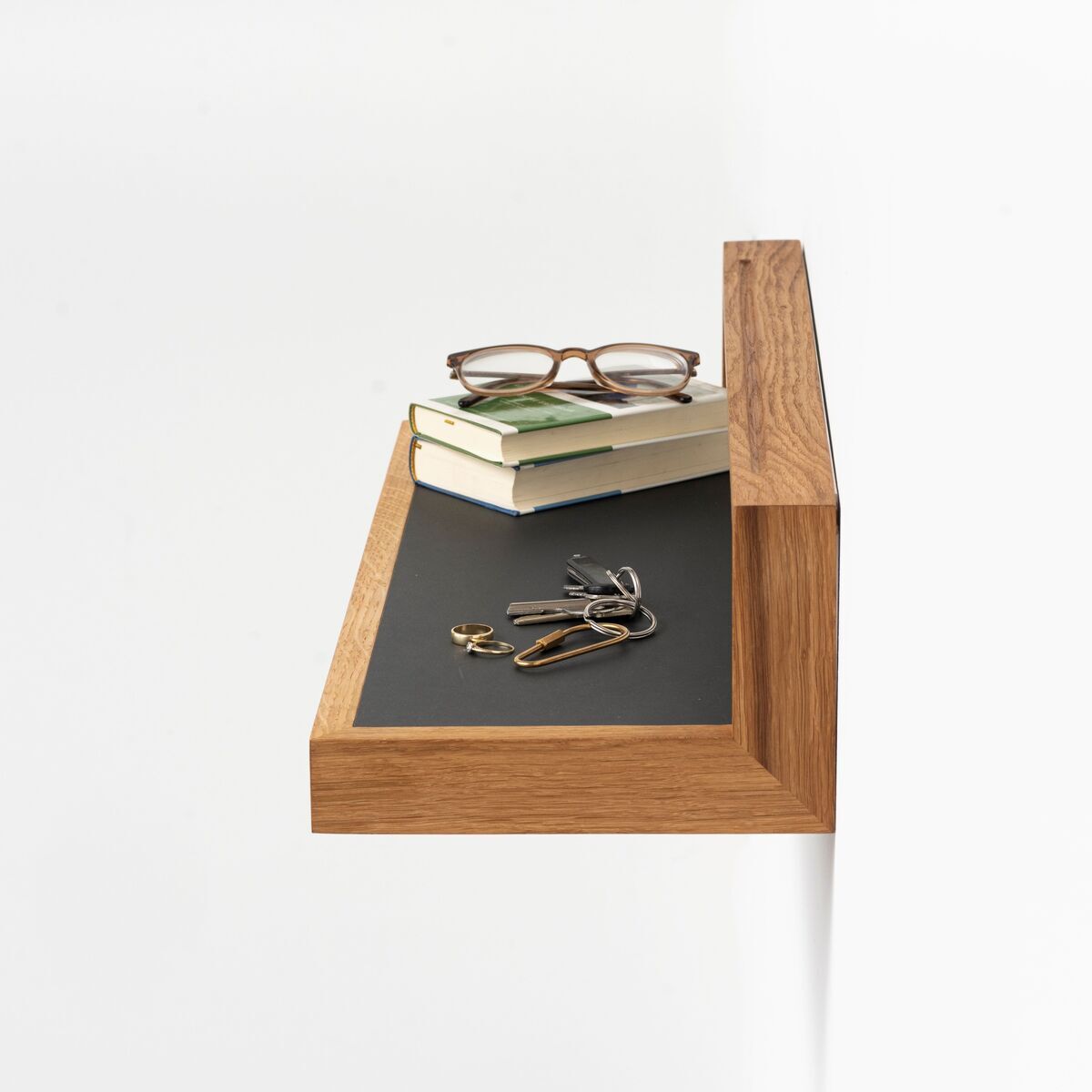 Ein modernes Ablagebord in Anthrazit und Eiche an einer weißen Wand montiert, bestückt mit Brillen, Büchern und einem Schlüsselbund.