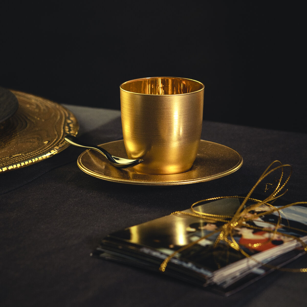 Ein goldfarbener Espressobecher mit passender Untertasse und Löffel steht auf einem dunklen Tisch neben einer dekorativ verschnürten Karten- oder Fotomappe. Elegantes und luxuriöses Ambiente, geeignet für festliche Anlässe oder als stilvolles Geschenk.