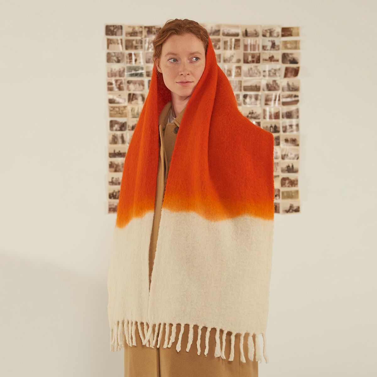 Eine Frau trägt den Schal »No.698« von MOISMONT in der Farbvariante tomato. Der Schal ist handgefertigt aus hochwertiger Schurwolle und Nylon, besitzt einen kräftigen dunkelorangen oberen Bereich, der in ein natürliches Weiß übergeht. Die Enden zieren feine Fransen. Im Hintergrund sind zahlreiche kleine, alte Fotos zu sehen.
