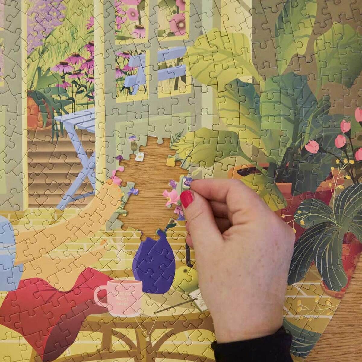 Eine Hand setzt ein Teil in ein fast fertiges 1000-Teile-Puzzle »Morning Glow« von VISSEVASSE ein. Das Motiv zeigt ein sonniges Interieur mit grünen Zimmerpflanzen, bunten Blumentöpfen, einer violetten Vase, einer rosa Tasse und großen Fenstern im Hintergrund.