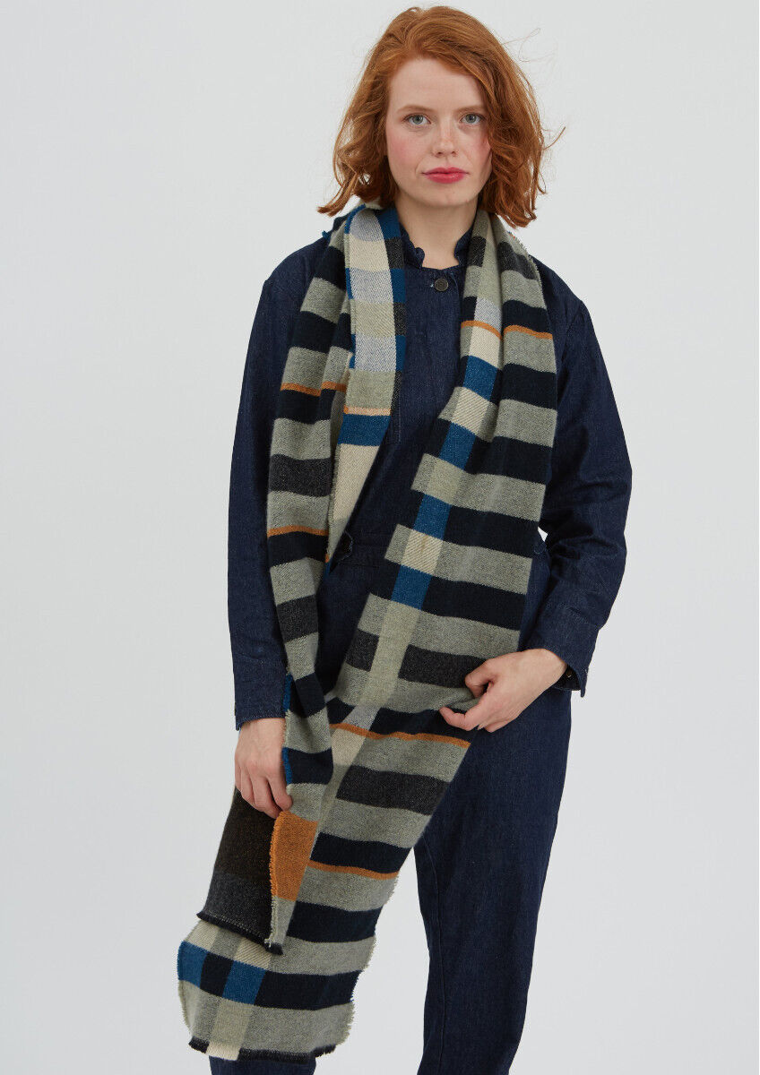 Eine Frau trägt einen langen Stölzl Schal im Bauhaus Design. Der Schal ist schwarz, grau, blau und orange gestreift. Sie trägt einen dunkelblauen Overall und hat mittellange, rote Haare.