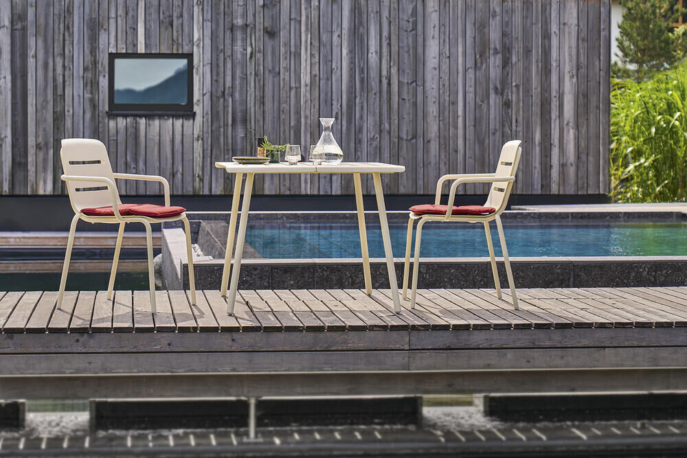 Zwei beige Sessel »VAPIO« an einem weißen Tisch auf einer Terrasse vor einem Pool und einer Holzhausfassade. Die Sessel haben rote Kissen und stehen auf einem hölzernen Terrassenboden.