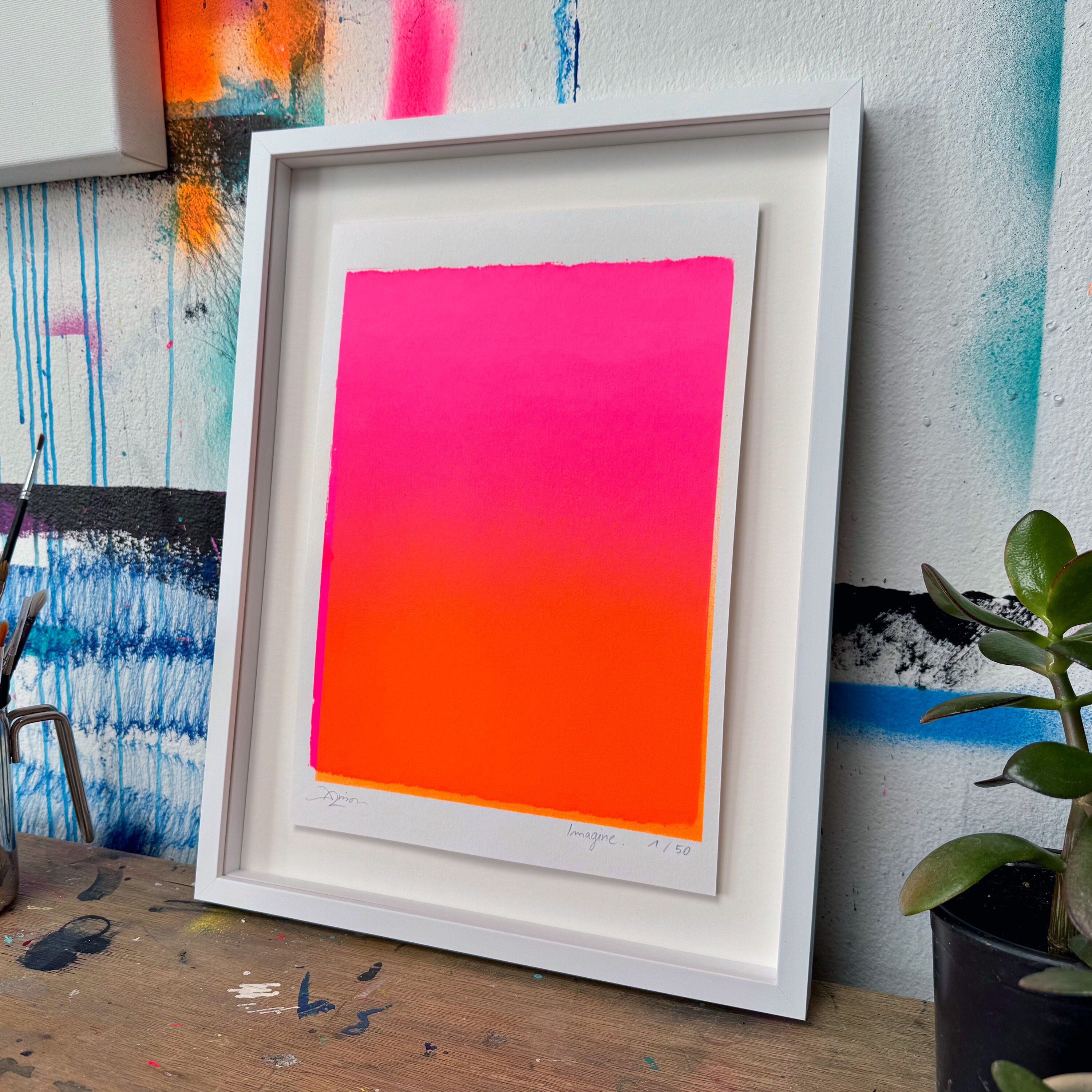 Abstraktes Kunstwerk in kräftigen Pink- und Orangetönen, mittig in weißem Rahmen präsentiert. Der Druck steht auf einem Schreibtisch vor einer Wand mit Farbspritzern und Pinselstrichen. Rechts im Vordergrund befindet sich eine kleine Topfpflanze. Das Bild vermittelt einen modernen, kreativen und farbenfrohen Eindruck als Wohn- oder Bürodekoration.
